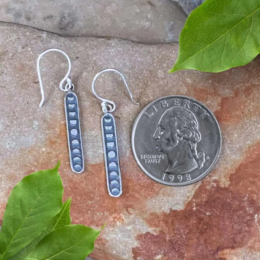 an image of a pair of Sterling Moon Phases Earrings shaped in a long vertical oval with the phases of the moon next to a quarter