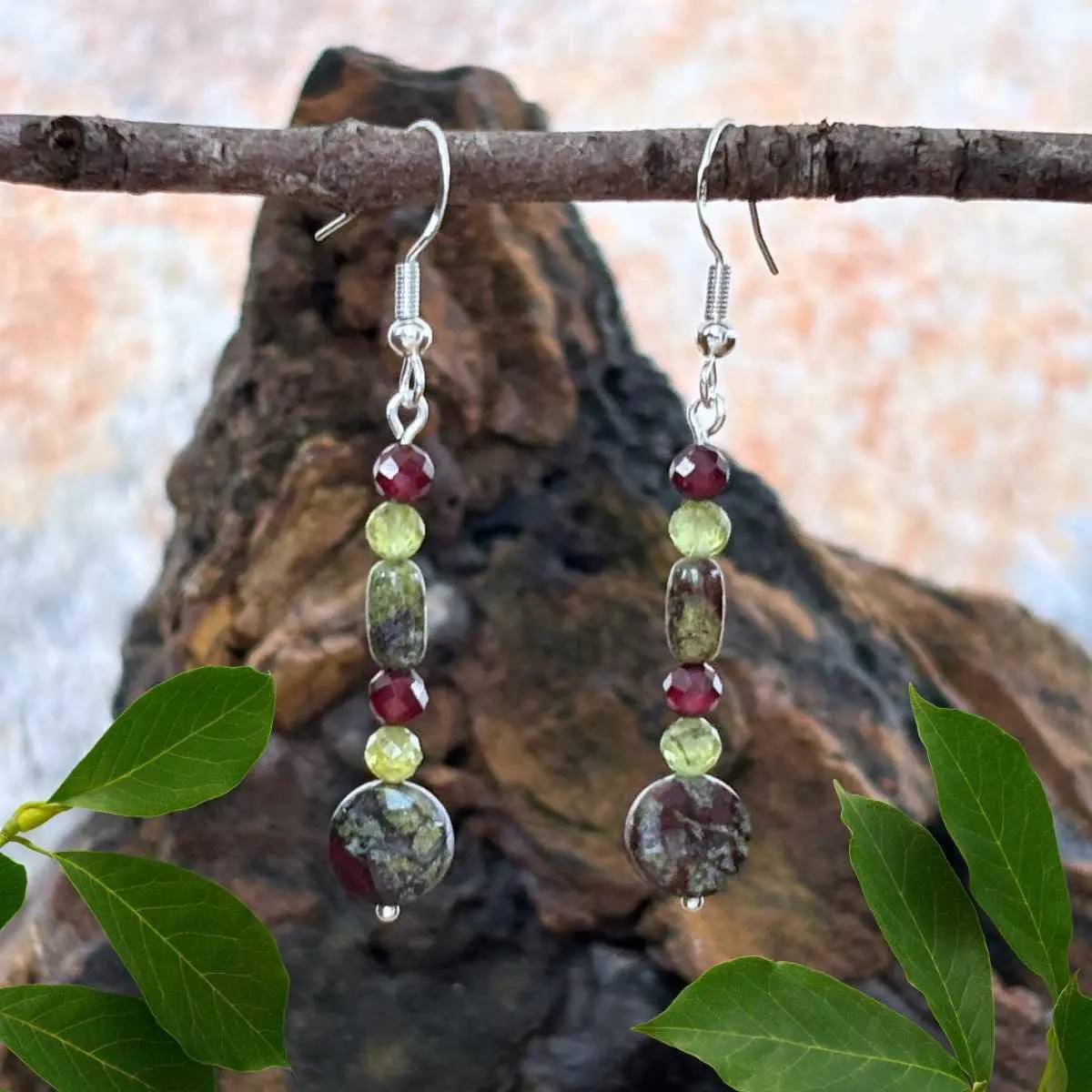 an image of a pair of Dragon Blood Jasper with Peridot and Garnet Earrings featuring a faceted coin shaped dragon blood jasper bead at the bottom, with a small faceted peridot and garnet bead and then a small smooth barrel shaped dragon blood jasper and topped off with another peridot and garnet bead. They are hanging on a sterling silver earring hook and have a medium dangle.