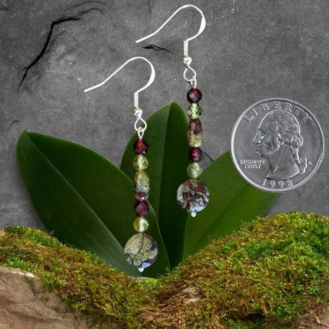 an image of a pair of Dragon Blood Jasper with Peridot and Garnet Earrings featuring a faceted coin shaped dragon blood jasper bead at the bottom, with a small faceted peridot and garnet bead and then a small smooth barrel shaped dragon blood jasper and topped off with another peridot and garnet bead. They are hanging on a sterling silver earring hook and have a medium dangle. The earrings are next to a quarter