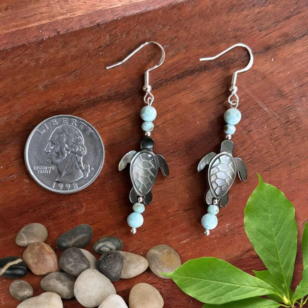 An image of a pair of Larimar and Black Mother of Pearl Sea Turtle Earrings. The earrings have a round smooth larimar bead and a smaller faceted larimar bead with a hand carved Black Mother of Pearl Sea Turtle and another set of the same larimar beads on top. They are on a sterling silver earring hook