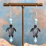 An image of a pair of Larimar and Black Mother of Pearl Sea Turtle Earrings. The earrings have a round smooth larimar bead and a smaller faceted larimar bead with a hand carved Black Mother of Pearl Sea Turtle and another set of the same larimar beads on top. They are on a sterling silver earring hook