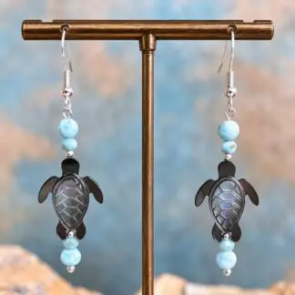 An image of a pair of Larimar and Black Mother of Pearl Sea Turtle Earrings. The earrings have a round smooth larimar bead and a smaller faceted larimar bead with a hand carved Black Mother of Pearl Sea Turtle and another set of the same larimar beads on top. They are on a sterling silver earring hook
