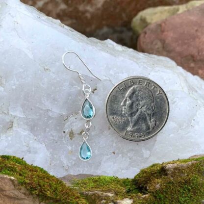 A photo of sterling silver hook dangle earrings with two dainty faceted blue topaz gemstones on each earrings in teardrop shape next to quarter for perspective