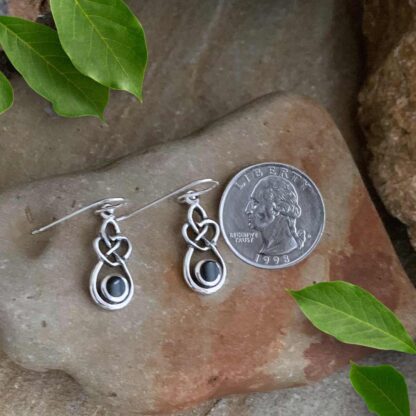 An image of a pair of Sterling Silver and Black Agate Celtic Heart Dangle Earrings with a total drop of .78 inches. The earrings are laying flat on a stone with a quarter next to them