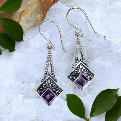 an image of an elegant pair of sterling silver dangle earrings that have two pieces of sterling silver dropping down to a square diamond shape with oxidation and a bit of a small swirl accent design and then a faceted square amethyst at the base angled in a diamond shape.