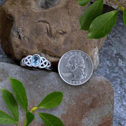 an image of our Sterling Silver and Oval Blue Topaz Celtic Ring. An oval faceted gemstone in the center with a Celtic knot on each side of the stone. The ring is next to a quarter for perspective