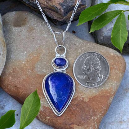 an image of a teardrop shaped lapis lazuli pendant with a smaller oval lapis lazuli stone above it, set in sterling silver. The pendant has a total drop of 2.5 inches and a width of .75 inches. The pendant is next to a quarter.