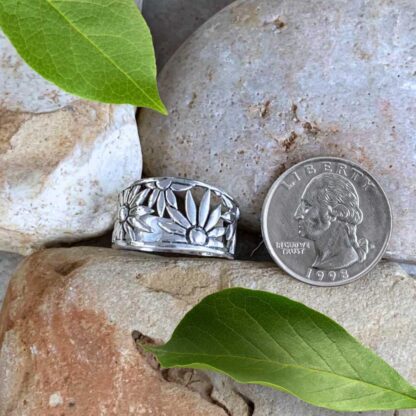 an image of a wide sterling silver ring with daisies wrapping around as the design tapers toward the back. Ring is next to a quarter