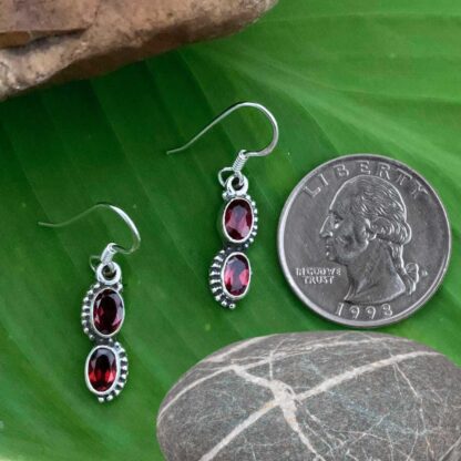 an image of a pair of dangle earrings with two oval faceted garnet gemstones and set in sterling silver. The earrings are next to a quarter