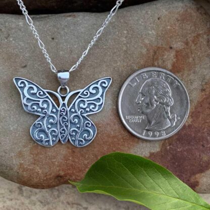 an image of a sterling silver butterfly pendant with oxidation and a swirling pattern all over next to a quarter. The pendant has a .866 total drop