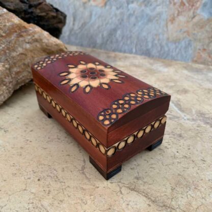 an image of a side angle of a small handcrafted brown trunk keepsake box with a floral design on the top and a circular pattern on the ends and around the sides. The dimensions are Length: 4.188 inches x Width: 2.125 inches x Height: 2 inches