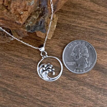 an image of a sterling silver round open pendant with ocean waves cresting on the left side of it. The pendant is next to a quarter