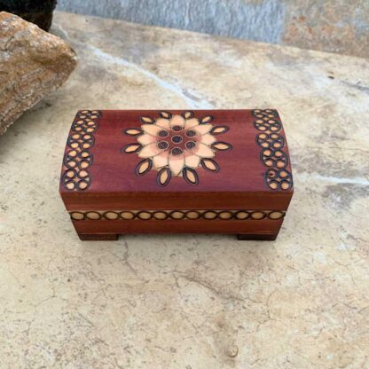 an image of a small handcrafted brown trunk keepsake box with a floral design on the top and a circular pattern on the ends and around the sides. The dimensions are Length: 4.188 inches x Width: 2.125 inches x Height: 2 inches