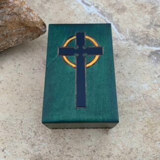 an image of a forest green handcrafted box with a black cross and a yellow circle on top and detailed trim around the bottom wrapping around the edge. It has a red velvet lined inside bottom. Dimensions are 4 x 2.38 x 1.50 inches.