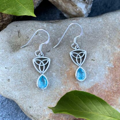 an image of a pari of sterling silver dangle earrings with a Celtic Trinity or Triquetra knot at the top and a teardrop shaped faceted blue topaz dangling from the bottom.