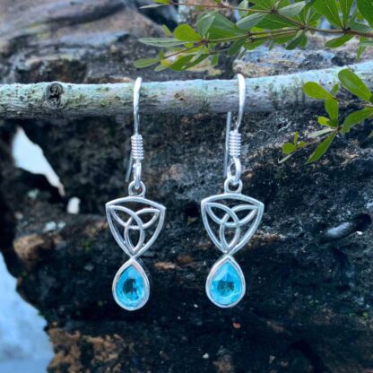 an image of a pari of sterling silver dangle earrings with a Celtic Trinity or Triquetra knot at the top and a teardrop shaped faceted blue topaz dangling from the bottom.