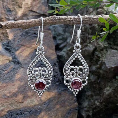 an image of a pair of sterling silver earrings with an upside down open heart and silver beadwork and a small round faceted garnet at the bottom.