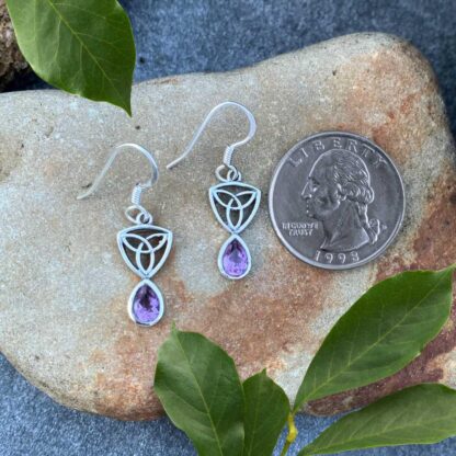 an image of a pari of sterling silver dangle earrings with a Celtic Trinity or Triquetra knot at the top and a teardrop shaped faceted amethyst dangling from the bottom. The earrings are next to a quarter