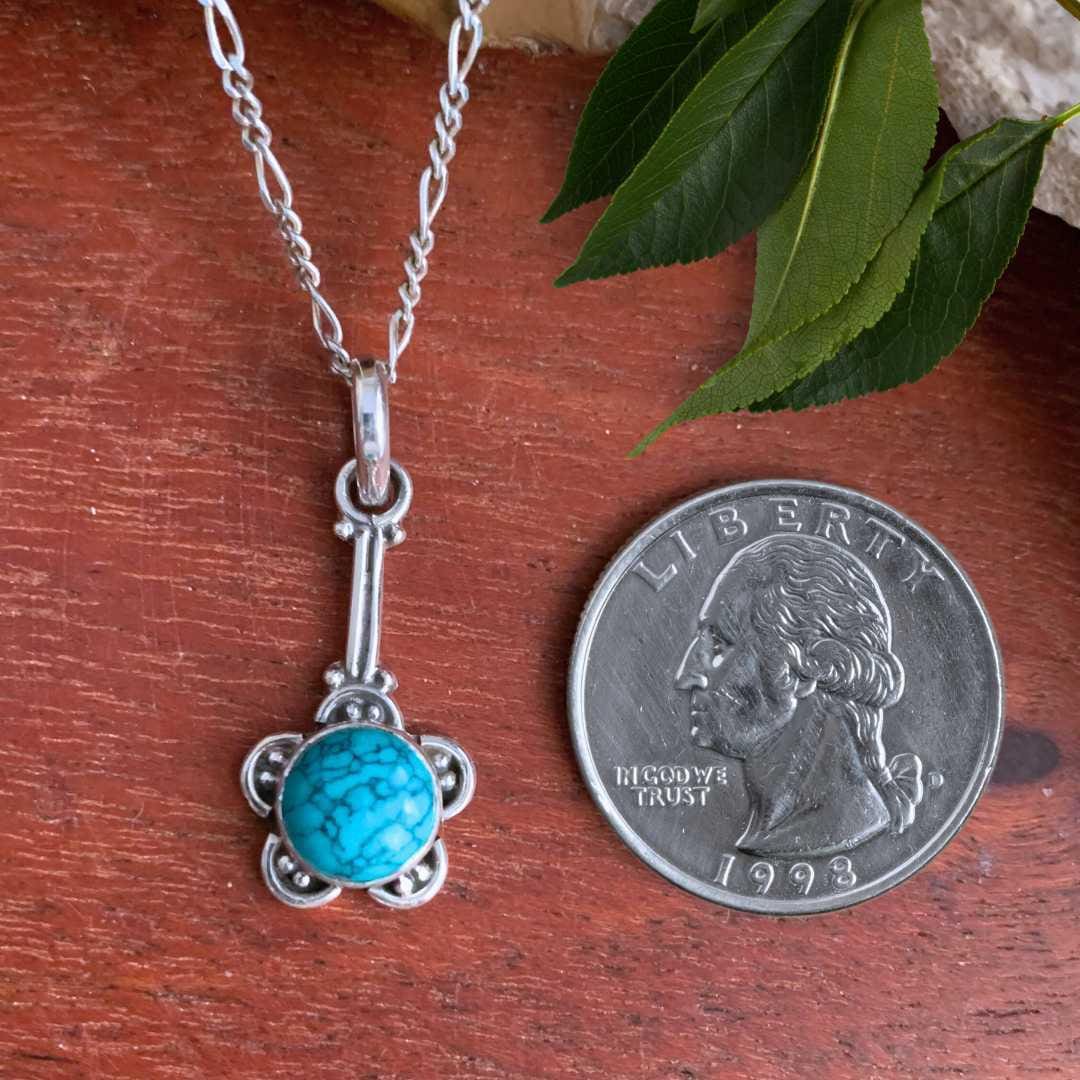 An image of a round turquoise stone surrounded a sterling silver flower shape handing from a sterling silver stem next to a quarter