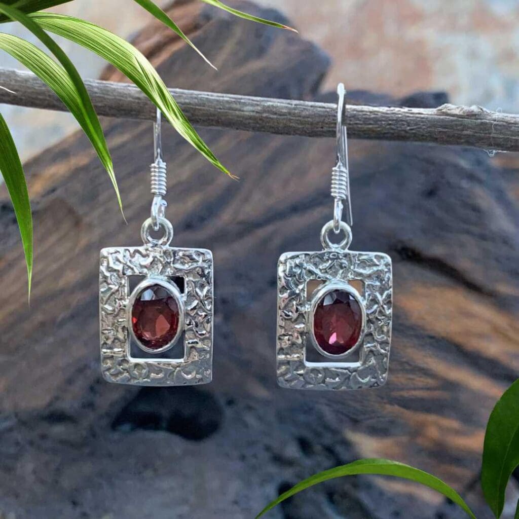 an image of a pair of Rectangular Sterling Silver and Garnet Earrings. They are textured sterling with an opening in the center and an oval faceted garnet gemstone.