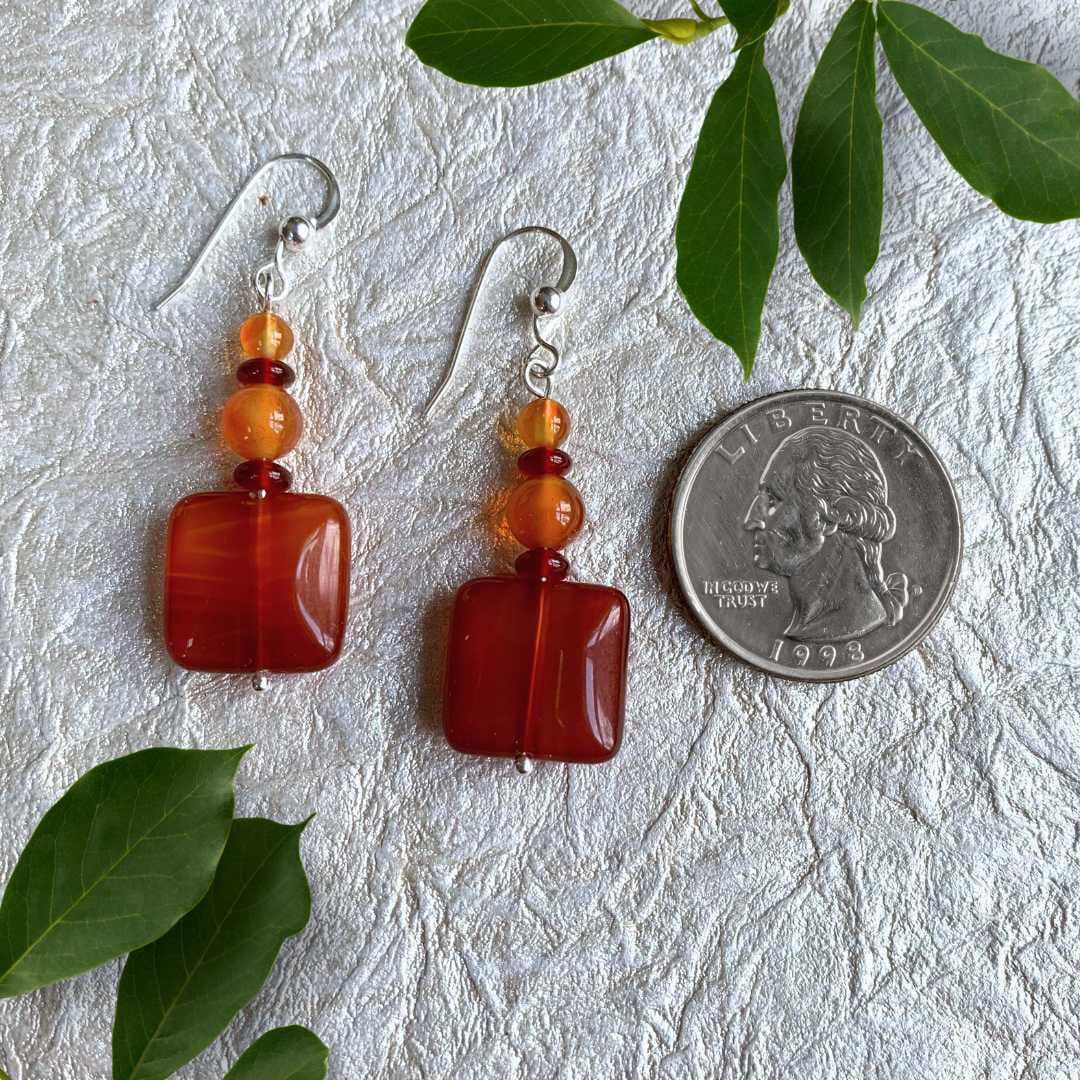 an image of a pair of Square Carnelian Mixed Beaded Earrings with a square carnelian gemstone at the bottom and alternating small rondelle and round beads going up above it. The earrings are next to a quarter for size perspective