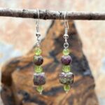 an image of a pair of Atlantisite Stichtite and Peridot Sterling Earrings featuring a square faceted cube bead of Atlantistite Stichtite with a smaller faceted peridot cube bead and a round smooth Atlantistite Stichtite bead. These are all sandwiched between a round faceted peridot 4 mm bead on the top and bottom and are on a sterling silver earring hook with medium dangle