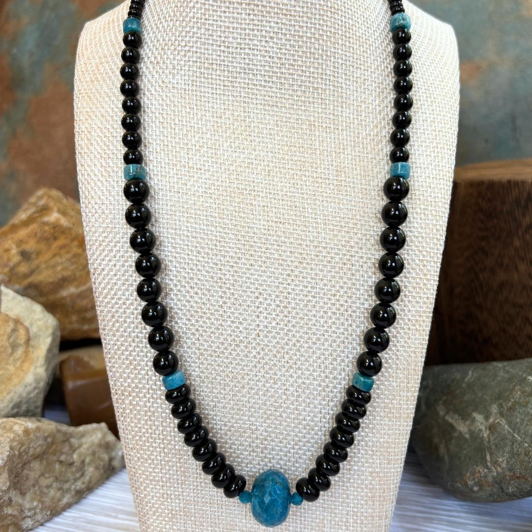 an image of an Apatite and Black Onyx Necklace featuring one large faceted rondelle of blue apatite in the center and rondelle and round black onyx beads working up the neck with occasional apatite heishi beads interspersed.
