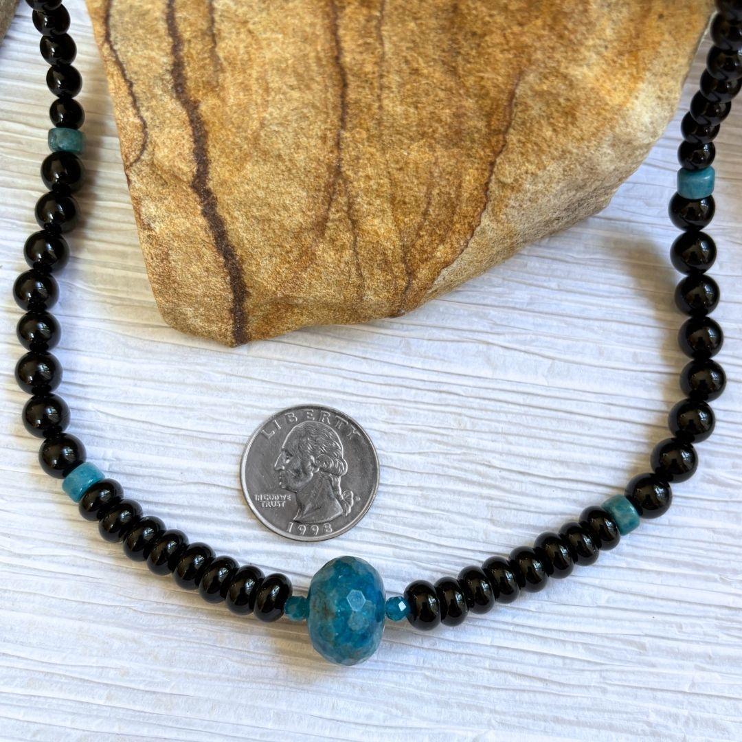 an image of an Apatite and Black Onyx Necklace featuring one large faceted rondelle of blue apatite in the center and rondelle and round black onyx beads working up the neck with occasional apatite heishi beads interspersed.