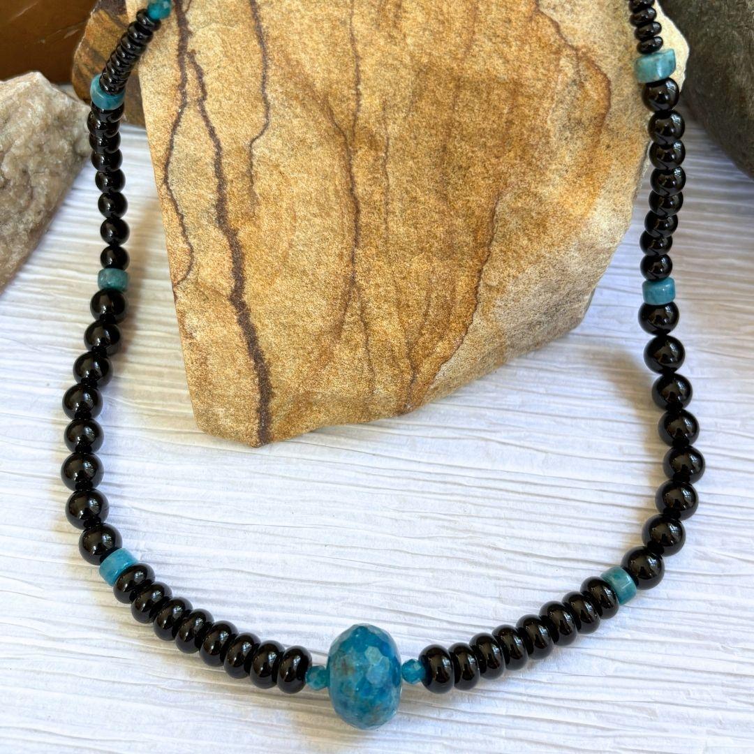 an image of an Apatite and Black Onyx Necklace featuring one large faceted rondelle of blue apatite in the center and rondelle and round black onyx beads working up the neck with occasional apatite heishi beads interspersed.
